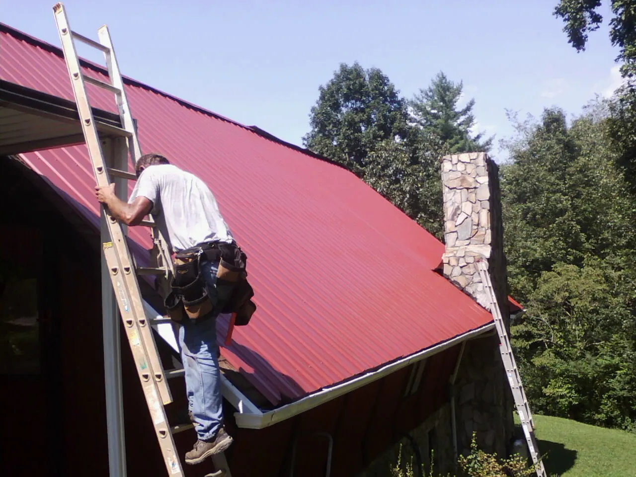 Roofer on a ladder beside a red metal roof and stone chimney — Soffit & Fascia Repair by Roofing Brothers in Springtown